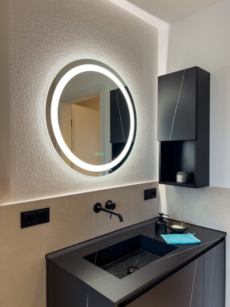 Modernes Badezimmer mit beleuchtetem, rundem Spiegel über einem schwarzen Waschtisch.