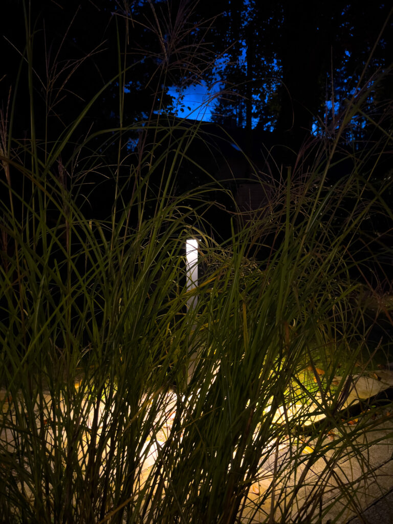 Moderne Außenstehleuchte beleuchtet sanft einen Gartenweg zwischen hohen Gräsern in der Dämmerung.