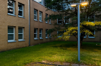 Moderne Mastleuchte beleuchtet Grünfläche und Parkplatz vor Bürogebäude in der Abenddämmerung