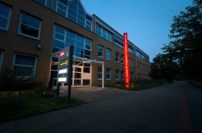 Architektonische Außenbeleuchtung eines Bürogebäudes mit beleuchtetem Eingangsbereich und vertikalem Lichtstelen-Element bei Dämmerung