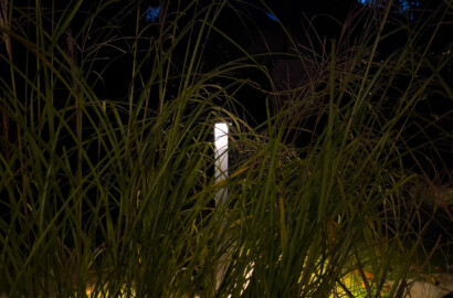 Moderne Außenstehleuchte beleuchtet sanft einen Gartenweg zwischen hohen Gräsern in der Dämmerung.