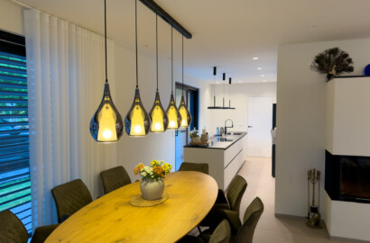 Moderne Lichtplanung in einer offenen Wohnküche mit einer Fünfer-Gruppe tropfenförmiger Pendelleuchten über dem ovalen Holz-Esstisch und dezenten Einbauspots im Küchenbereich.