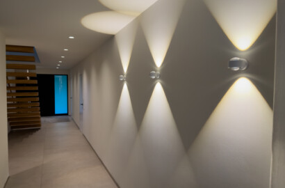 Moderner Flur mit professioneller Lichtplanung durch Up-&-Down-Wandleuchten und dezent integrierte LED-Einbauspots. Die Lichtinszenierung erzeugt geometrische Lichtkegel und betont die klare Architektur sowie die geradlinige Raumgestaltung.
