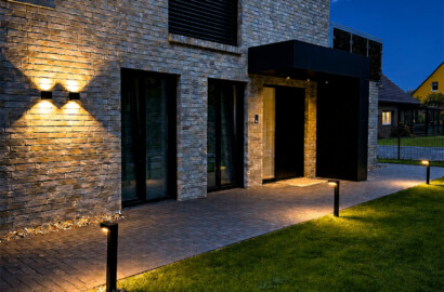 Moderne Außenbeleuchtung eines Einfamilienhauses bei Nacht mit minimalistischen Pollerleuchten entlang des gepflasterten Weges und Up-and-Down-Wandstrahlern an der Klinkerfassade.