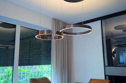 Nahaufnahme von zwei ringförmigen Pendelleuchten in edlem Metallic-Finish mit integrierten LED-Bändern über einem dunklen Esstisch.