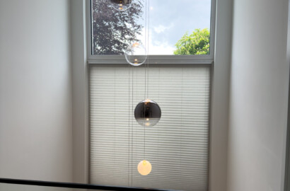 Blick von einer Galerie hinab in einen Luftraum mit einer kaskadenförmigen Pendelleuchte aus spiegelnden, transparenten und opalen Glaskugeln vor einer großen Fensterfront.