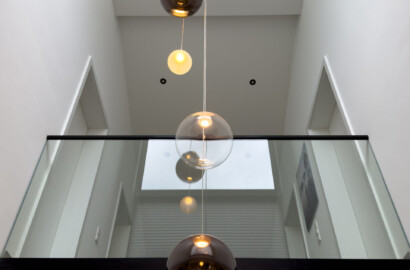 Blick von unten in einen offenen Luftraum mit einer modernen Kaskaden-Pendelleuchte aus verschiedenen großen, transparenten und getönten Glaskugeln vor einer Glasgalerie.