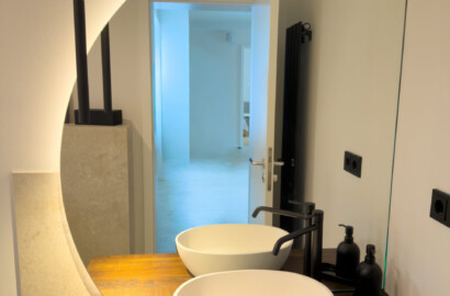 Moderne Badezimmer-Lichtplanung mit indirekter LED-Hintergrundbeleuchtung an einem organisch geformten Wandspiegel und Holzwaschtisch.