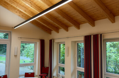 Gruppenraum mit Tischen und einer abgependelten schwarzen LED-Linearleuchte unter der Holzdecke