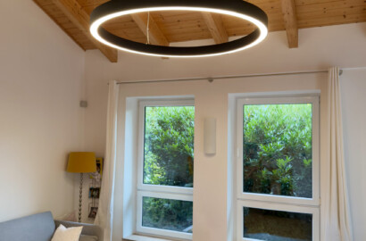 Schwarze LED-Ringpendelleuchte unter einer Holzdecke in einem Raum mit Sofa und großen Fenstern