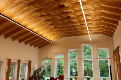 Großer Saal mit Holzdachstuhl und zwei LED-Linearpendelleuchten, die die Holzdecke in warmweißem Licht betonen