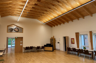 Veranstaltungssaal mit zwei LED-Linearpendelleuchten unter der Holzdecke, im Hintergrund Klavier und Giebelfenster